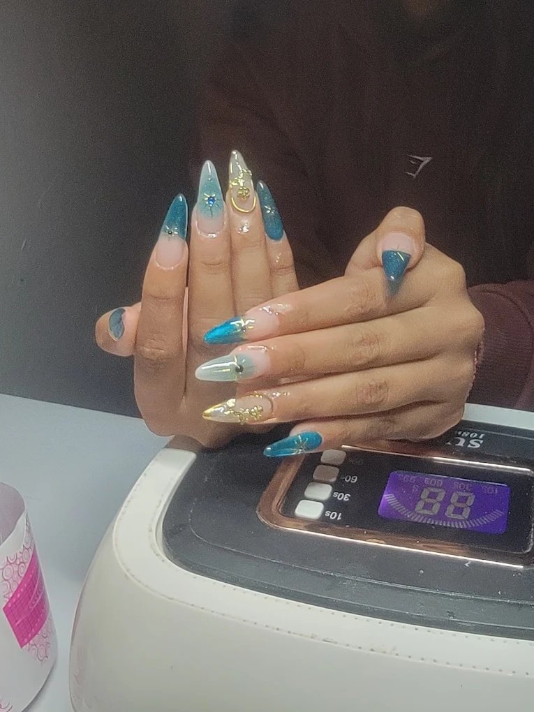 Manicura en stiletto larga con efectos holográficos azules y detalles de joyería de metal dorado